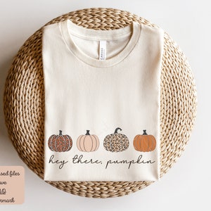 May include: A beige t-shirt with the text "hey there, pumpkin" and four pumpkins in a row. The pumpkins are orange, peach, and leopard print.
