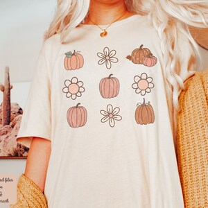 Op de afbeelding: Een wit T-shirt met een patroon van roze en bruine pompoenen en bloemen. De pompoenen hebben stelen en bladeren. De bloemen hebben vijf bloemblaadjes.