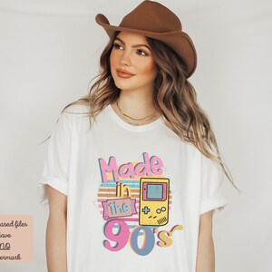 May include: White t-shirt with a graphic design that says "Made in the 90's" with a retro video game controller and a sunset behind it.