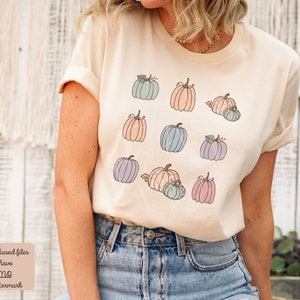 May include: A beige t-shirt with a pattern of pastel-colored pumpkins. The pumpkins are in various sizes and have a simple, hand-drawn style.