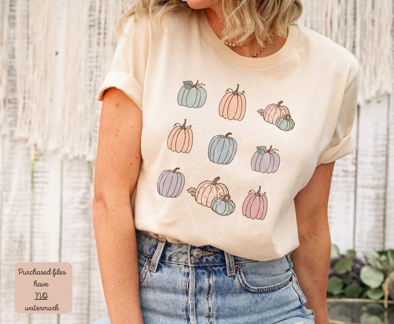 May include: A beige t-shirt with a pattern of pastel-colored pumpkins. The pumpkins are in various sizes and have a simple, hand-drawn style.