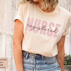 Puede incluir: Una camiseta beige con el texto "NURSE registered" en rosa con un aspecto desgastado.