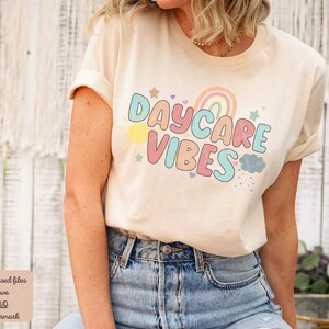 May include: A cream-colored t-shirt with rolled-up sleeves featuring the text "DAYCARE VIBES" in colorful bubble letters. The design includes a rainbow, sun, stars, and clouds. The shirt is paired with blue jeans.