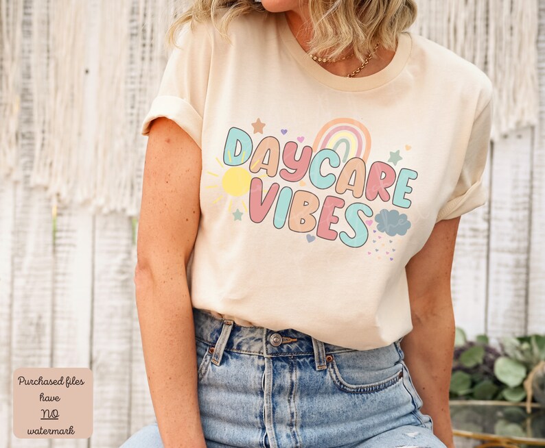 May include: A cream-colored t-shirt with rolled-up sleeves featuring the text "DAYCARE VIBES" in colorful bubble letters. The design includes a rainbow, sun, stars, and clouds. The shirt is paired with blue jeans.