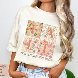 Op de afbeelding: Een wit T-shirt met een bloemenontwerp dat "FALL" in grote letters spelt. De tekst "FOR JESUS HE NEVER LEAVES" is onder het bloemenontwerp gedrukt.