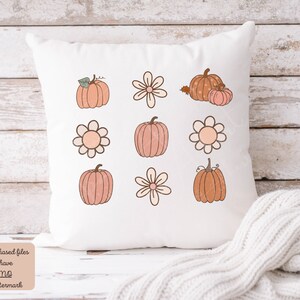 Op de afbeelding: Een wit kussen met een ontwerp van negen herfstthema-illustraties. De illustraties omvatten pompoenen en bloemen in tinten oranje, roze en bruin.