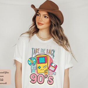 K&ouml;nnte beinhalten: Wei&szlig;es T-Shirt mit einem Grafikdesign, das ein Retro-Videospiel, eine Diskette und eine CD zeigt. Der Text auf dem Shirt lautet "Take Me Back to the 90's".