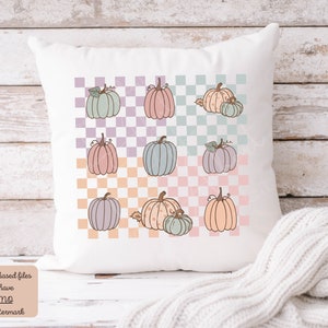 May include: A white cushion with a checkered pattern in pastel colours. The pattern features a repeating design of pumpkins in various shades of pink, blue, and orange.