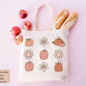 Op de afbeelding: Een witte tote bag met een ontwerp van pompoenen en bloemen. De tas ligt op een roze oppervlak met appels en stokbroden.