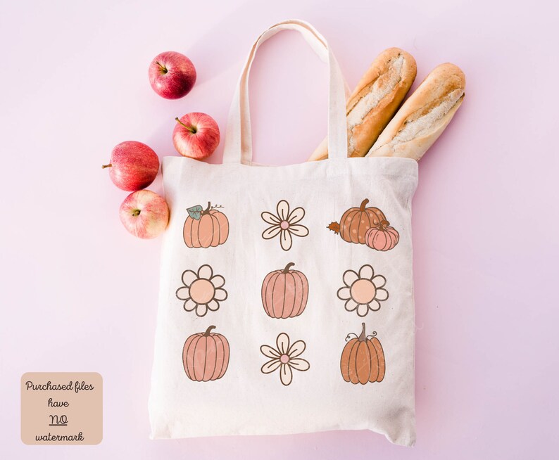 Op de afbeelding: Een witte tote bag met een ontwerp van pompoenen en bloemen. De tas ligt op een roze oppervlak met appels en stokbroden.