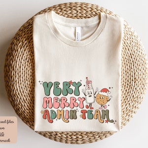 May include: A beige t-shirt with a Christmas-themed graphic that says "Very Merry Admin Team". The graphic features a cartoon milk carton and a cookie with Santa hats.