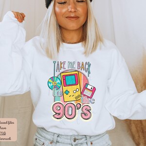 K&ouml;nnte beinhalten: Wei&szlig;er Pullover mit einem bunten Grafikdesign, auf dem "Take me back to the 90's" steht. Die Grafik enth&auml;lt ein Kassettenband, eine Videospielkonsole und eine Diskette.