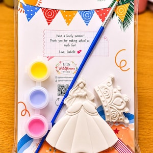 Könnte beinhalten: Ein Bastelset in einer transparenten Kunststoffhülle mit einer weißen Prinzessinnenfigur, einer weißen Kronenfigur, einem blauen Pinsel und drei kleinen Farbtöpfen in Gelb, Hellblau und Pink auf einer sommerlich gestalteten Karte.