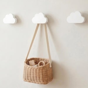 May include: Three white cloud-shaped wall hooks mounted on a plain wall. One hook supports a small, light brown woven wicker basket with a flat strap handle. The basket contains a few wooden blocks and a ring-shaped toy inside the open top.