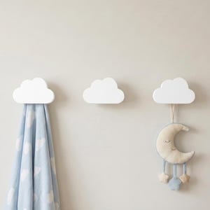 Puede incluir: Tres ganchos de pared blancos con forma de nube montados horizontalmente. Una manta azul claro con un patrón de nubes blancas cuelga del gancho izquierdo, y un móvil de peluche de media luna con estrellas cuelga del gancho derecho.