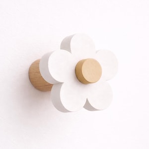 May include: A white flower-shaped wall hook with a light brown centre and a wooden base. The hook is mounted on a white wall. The flower petals are rounded and painted white, creating a simple, modern design.