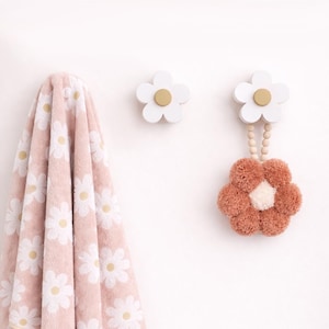 May include: A pink daisy-patterned towel hangs next to two white flower-shaped wall hooks with gold centers. One hook holds a decorative pom-pom flower. The items are against a white wall, creating a clean, floral-themed display.