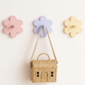 May include: Three flower-shaped wall hooks in pastel pink, light purple, and pale yellow. A small, rectangular, woven wicker basket with a handle and a house-like design featuring a door and two windows hangs from the centre purple hook.