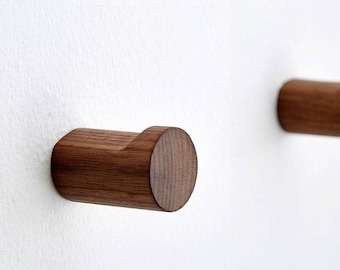Könnte beinhalten: Zwei Wandhaken aus Holz mit abgerundetem, zylindrischem Design. Die Haken sind aus dunkelbraunem Holz und an einer weißen Wand montiert.