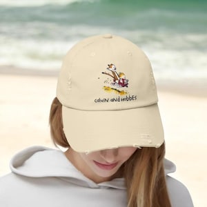 Op de afbeelding: Beige baseballpet met een gerafelde rand. De pet heeft een kleurrijke afbeelding van Calvin en Hobbes, met de tekst "calvin AND hobbes" eronder. De pet wordt door een persoon gedragen.