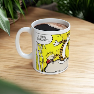 Könnte beinhalten: Ein weißer Keramikbecher mit einer farbigen Comic-Illustration an der Seite. Die Grafik zeigt einen kleinen Jungen und einen Tiger, die auf einem Felsen vor einem leuchtend gelben Hintergrund stehen, mit einer Sprechblase und dem Text I HATE EVERYBODY.
