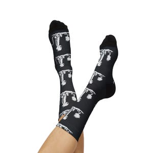 May include: Black socks with a white pattern of stylized, repeating figures. The socks have a black heel and toe, and the pattern covers the entire sock. The socks are knee-high and are shown on a person's legs.