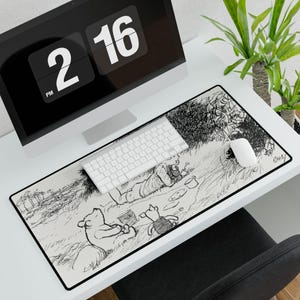 May include: A rectangular desk mat featuring a black and white line art illustration of Winnie the Pooh, Piglet, and Christopher Robin in a grassy field. The design includes a black border and the signature E.H. Shepard style sketch artwork.