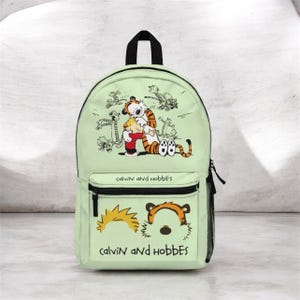 May include: A light green backpack featuring a graphic of Calvin and Hobbes hugging, with smaller sketches of the characters surrounding them. The front pocket displays the text Calvin and Hobbes above and below stylized outlines of their heads.