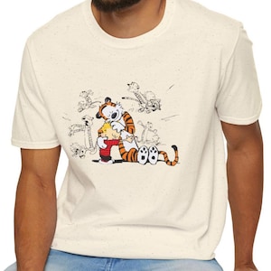Puede incluir: Camiseta color crema con una ilustración de dibujos animados de Calvin y Hobbes. El gráfico muestra a Calvin y Hobbes en una escena juguetona. La camiseta tiene una textura moteada y un cuello redondo clásico.