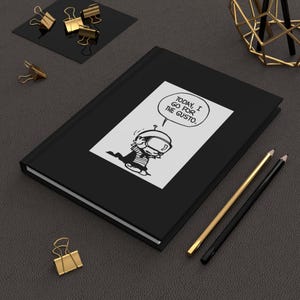 May include: A black hardcover notebook featuring a white rectangular graphic on the front cover. The graphic depicts a cartoon character wearing a helmet with the speech bubble text: TODAY, I GO FOR THE GUSTO. The notebook has a smooth, dark finish.