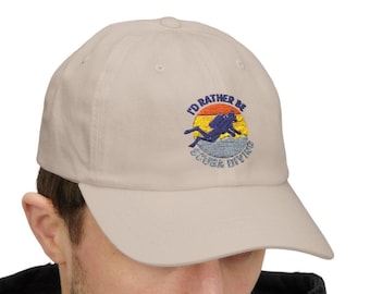 May include: A beige baseball cap with the embroidered text "I'd rather be scuba diving." The design features a diver silhouette in blue and yellow, set against a sunset backdrop. The cap is a classic, casual accessory.