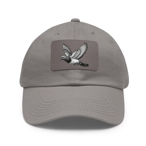 Puede incluir: Una gorra de béisbol de algodón gris con un parche que presenta una ilustración en blanco y negro de una paloma en vuelo.