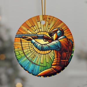 May include: Round ceramic ornament featuring a stylized illustration of a person aiming a shotgun. The design includes a colorful background with orange, yellow, green, and blue hues. The ornament has a gold string for hanging.
