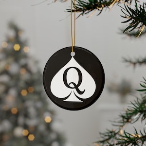 May include: A round black ceramic ornament featuring a white spade symbol with a large black letter Q centred inside. The ornament hangs from a thin gold string, providing a high-contrast design that stands out against the blurred holiday background.