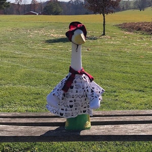 May include: A white goose figurine wearing a red and white patterned shirt and a red hat sits on a wooden bench in a field. The goose is looking directly at the camera.