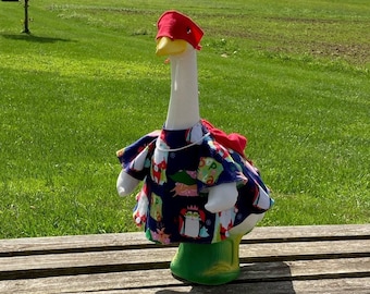 May include: A white goose figurine wearing a red hat and a colorful patterned dress. The goose is sitting on a wooden bench in front of a green grassy field.
