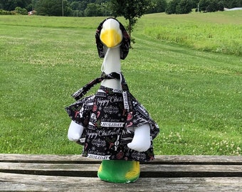 May include: A white goose figurine wearing a black and white holiday-themed outfit with a red and green hat. The goose is sitting on a wooden bench in a grassy field.