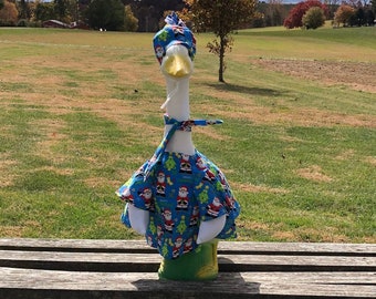 May include: A white goose wearing a blue and green Christmas-themed outfit is sitting on a wooden bench. The goose is facing the camera and has a yellow beak. The bench is made of wood and has a natural finish. The goose is sitting on a grassy field with a cloudy sky in the background.