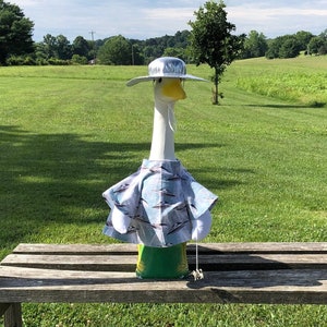 May include: A white goose figurine wearing a blue and white patterned shirt and a white hat. The goose is sitting on a wooden bench in a grassy field.