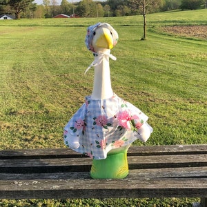 May include: A white goose figurine wearing a blue and white gingham dress with pink flowers. The goose is sitting on a wooden bench outdoors.
