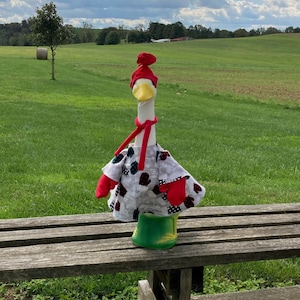 May include: A white goose wearing a red and white hat, a red scarf, and a gray and black patterned coat with red mittens. The goose is sitting on a wooden bench in a field.