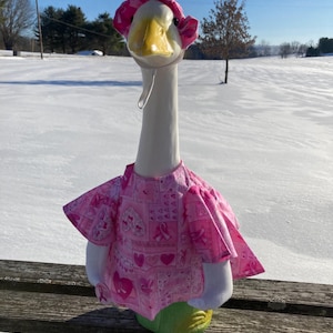 May include: A decorative goose figurine wearing a pink hat and a matching pink top with a breast cancer awareness ribbon pattern. The goose has a white neck and body, a yellow beak, and is standing on a green base.