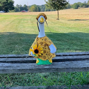May include: A goose figurine dressed in a yellow sunflower-patterned dress and hat, holding a sunflower. The goose has a white body, yellow beak, and black eyes. It stands on a green base, set against a backdrop of a wooden bench and a grassy field.