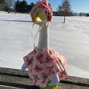 May include: A decorative goose figurine wearing a pink floral print dress and matching bonnet. The goose has a yellow beak and is set against a snowy winter landscape. The dress features a ribbon design.