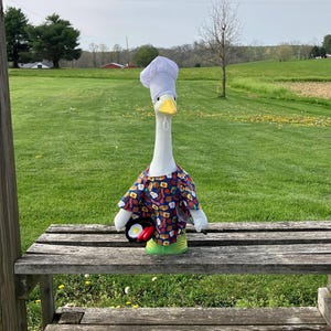 May include: A white goose wearing a chef's hat and a blue, red, and yellow shirt with a breakfast food pattern. The goose is holding a frying pan with a fried egg and a red sausage.