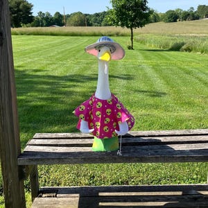 May include: A decorative goose figurine wearing a pink dress with yellow alien faces, a blue hat, and white gloves. The goose is standing on a wooden bench in a grassy outdoor setting.