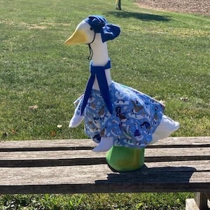 May include: A white goose figurine wearing a blue dress and hat with a winter scene pattern. The goose is standing on a wooden bench.