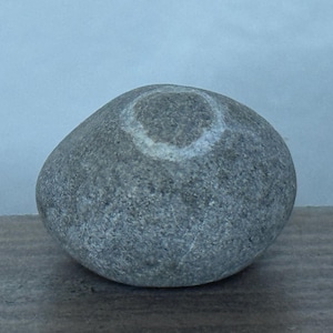 May include: A smooth, gray stone with a lighter-colored, circular marking on its surface. The stone is oval-shaped and sits on a wooden surface against a light blue background. The stone appears to be a natural, found object.