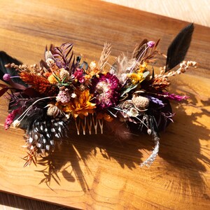 May include: A hair comb adorned with a dense arrangement of dried flowers and feathers. The composition features deep burgundy, orange, and yellow blossoms, accented by black feathers and white-spotted black feathers, all attached to a metal comb.