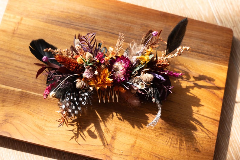 May include: A hair comb adorned with a dense arrangement of dried flowers and feathers. The composition features deep burgundy, orange, and yellow blossoms, accented by black feathers and white-spotted black feathers, all attached to a metal comb.